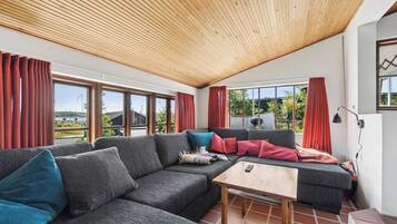 A modern living room with a wooden ceiling, large windows, and a sofa with multiple cushions.
