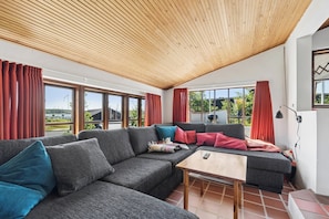 Ein modernes Wohnzimmer mit Holzboden, großen Fenstern und einem Sofa mit vielen Kissen.