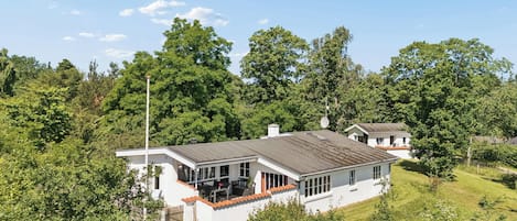 A white house with a grey roof, surrounded by lush greenery and trees.