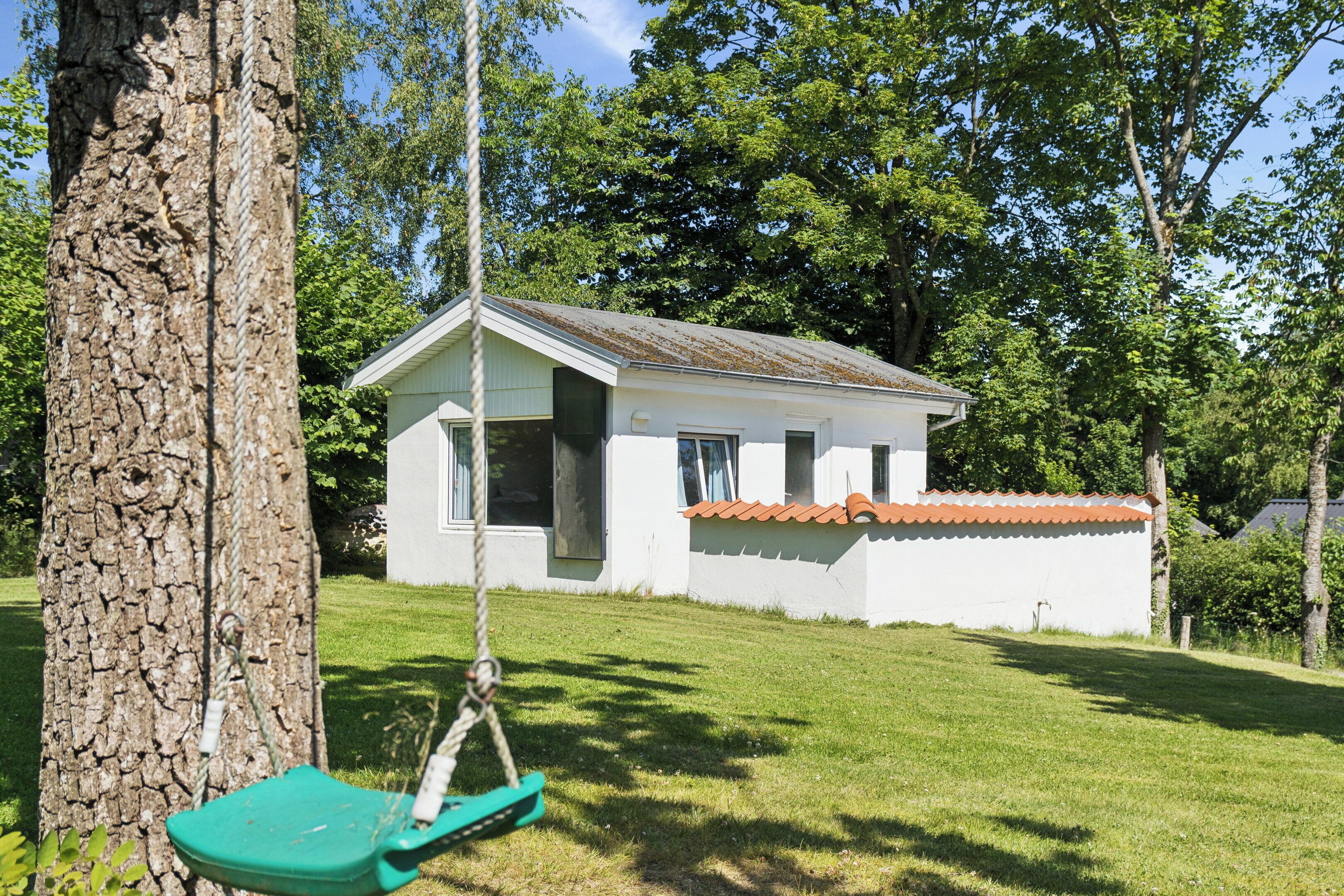 Une petite maison blanche avec un toit rouge, une balançoire et un arbre au premier plan.