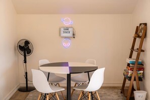 A room with a round table, two white chairs, a standing fan, and a neon sign on the wall. - Sun & Rally Retreat | Dual Swim Spa | Pickleball (Yucca Valley)