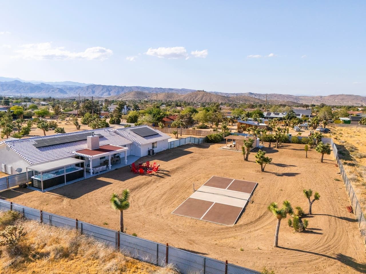 A large building with a swimming pool, tennis court, and fenced area with palm trees.