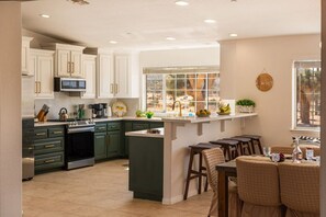 A kitchen with a central island, bar stools, a microwave, and a window with a view. - Sun & Rally Retreat | Dual Swim Spa | Pickleball (Yucca Valley)
