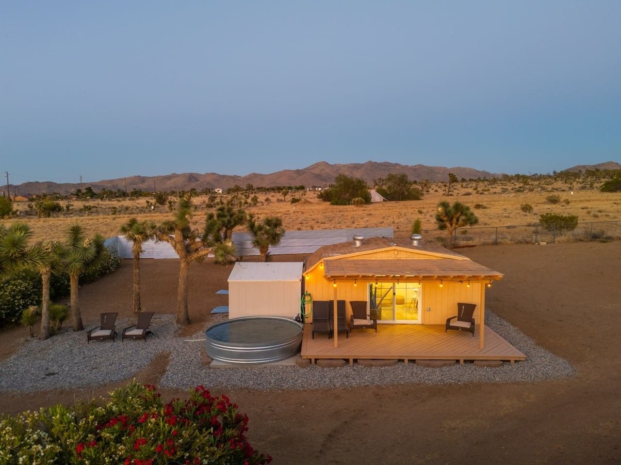 A desert-style cabin with a wooden deck, a hot tub, and a small pool, surrounded by sparse vegetation and hills in the background.