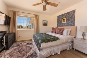 A bedroom with a bed, a ceiling fan, a TV, a desk, a lamp, and a window with curtains. - Sun & Rally Retreat | Dual Swim Spa | Pickleball (Yucca Valley)