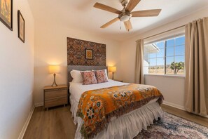 A bedroom with a bed, two bedside tables, a ceiling fan, a window with curtains, and a patterned wall. - Sun & Rally Retreat | Dual Swim Spa | Pickleball (Yucca Valley)