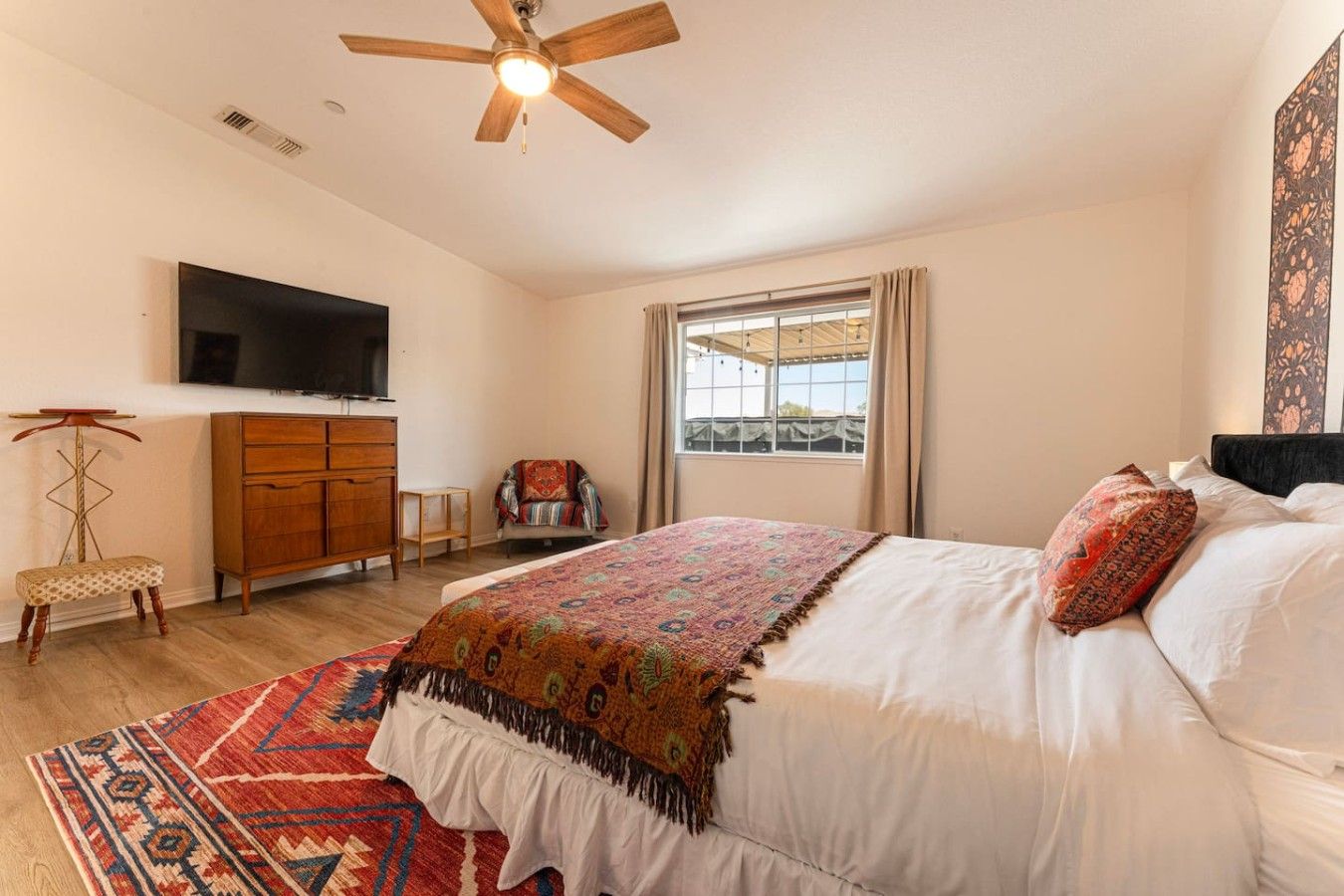 A bedroom with a bed, a television, a ceiling fan, a window with curtains, and a patterned rug.