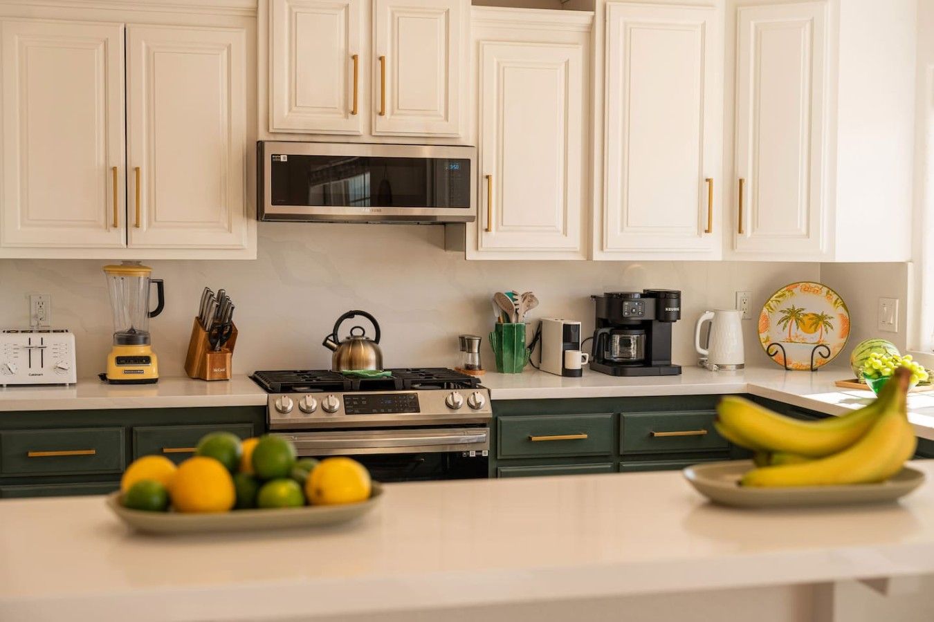 A kitchen with white cabinets, a stove, a microwave, and various appliances. There are fruits on the counter and a plate of lemons and limes.
