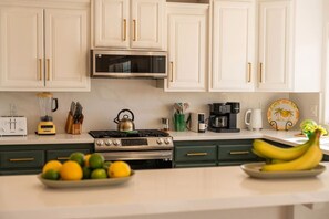 Une cuisine avec des meubles blancs, une cuisinière, un four à micro-ondes et divers appareils électroménagers. On y trouve des fruits sur le comptoir, ainsi qu’une assiette de citrons et de limes.