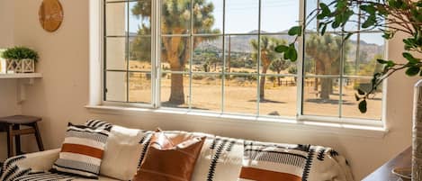 A room with a large window, a sofa with patterned cushions, a small table, and a potted plant.