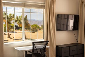 Una habitación con un ventanal grande, un escritorio, una silla, un televisor montado en la pared y un armario.