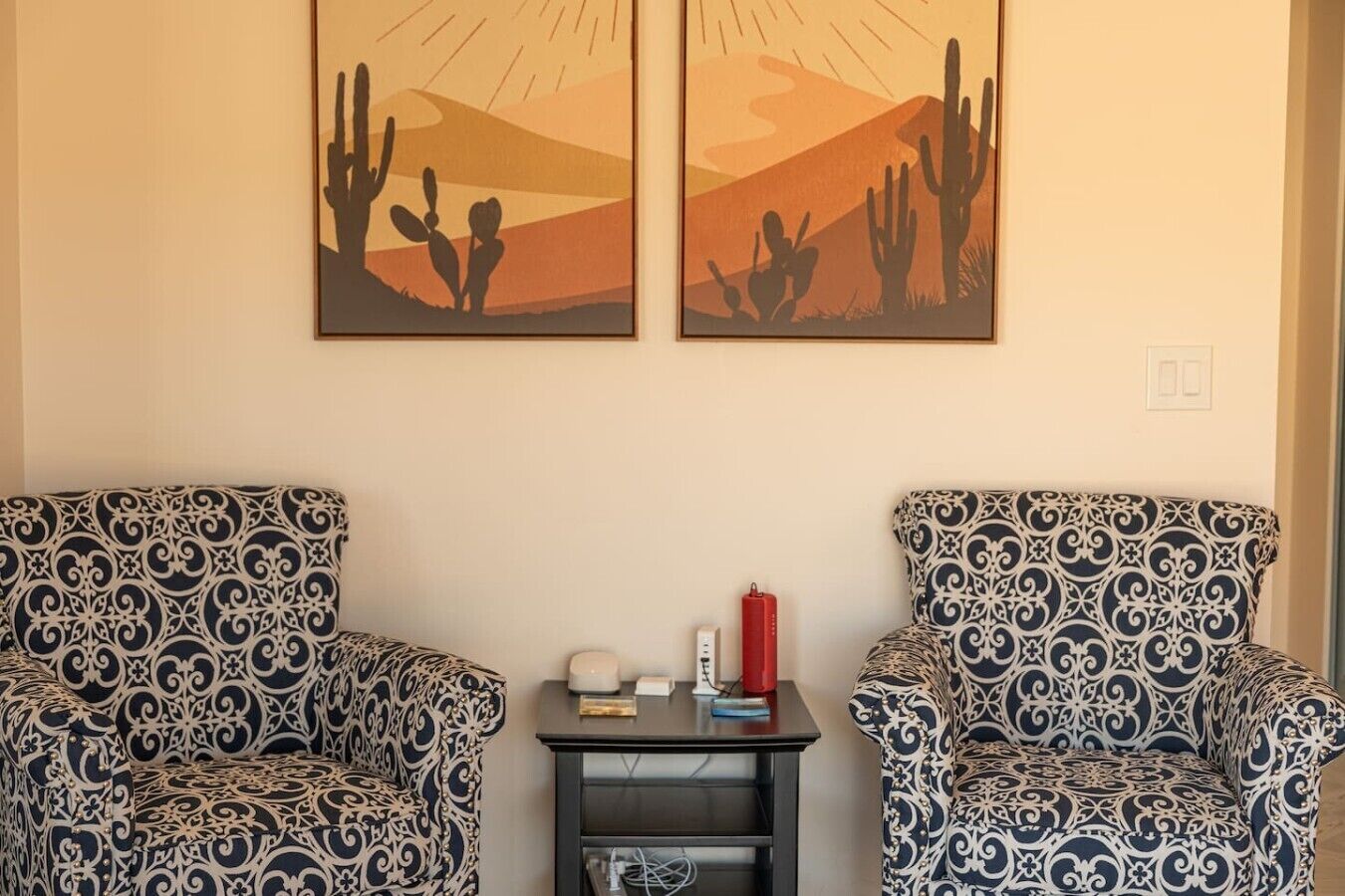A room with two patterned armchairs, a small table with a red lamp, and two wall-mounted paintings of cacti.