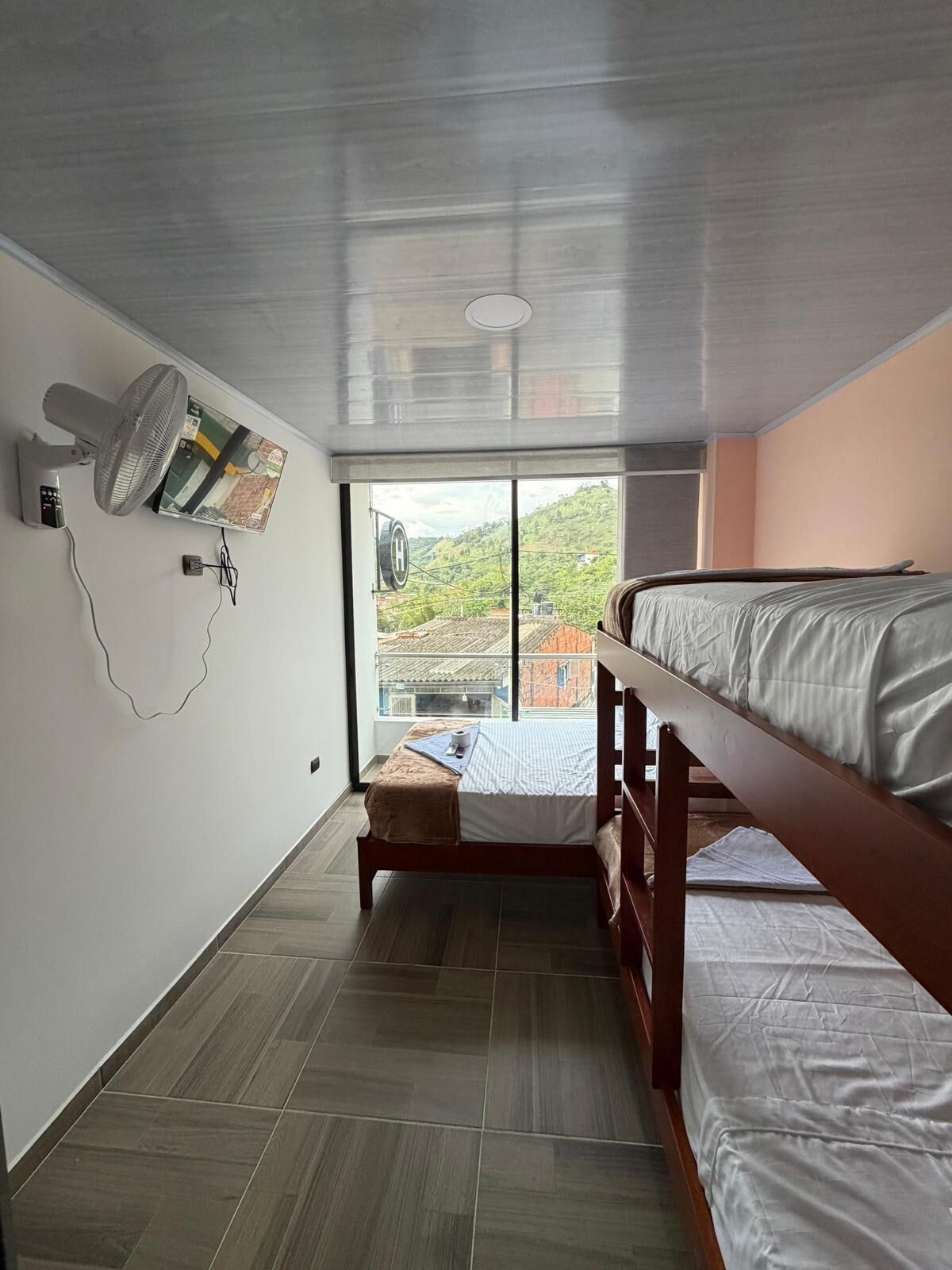 Una habitación con cama, ventilador de pie, ventana con vista a edificios y aire acondicionado instalado en la pared.