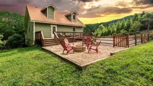 Ein grünes Haus mit einer Terrasse, zwei Adirondack-Stühlen, einer Feuerstelle und einem Holzzaun.