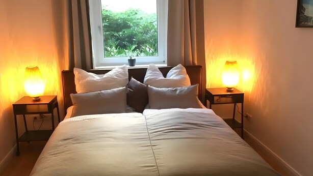 A bed with white bedding, two bedside lamps, a window with a view of greenery, and a small potted plant on the windowsill. - Apartment for 4 Guests, 55 m², with Wi-Fi (Berlin)