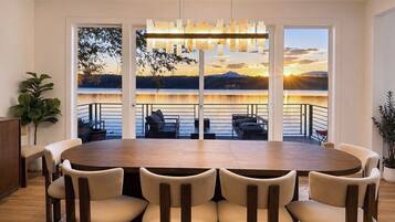 Un coin repas avec une grande table, six chaises et une vue sur un lac au coucher du soleil.