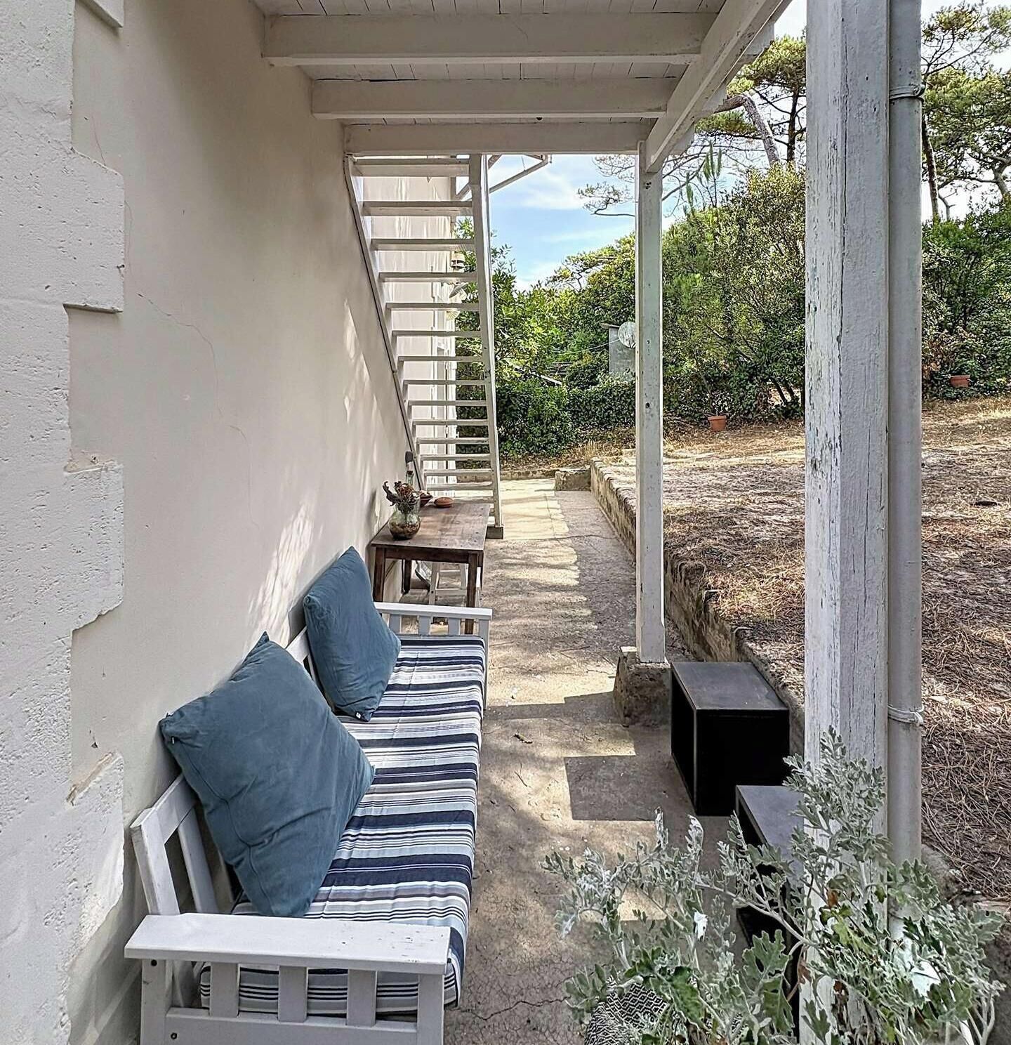 A covered outdoor seating area with a bench and a table, surrounded by plants and trees.