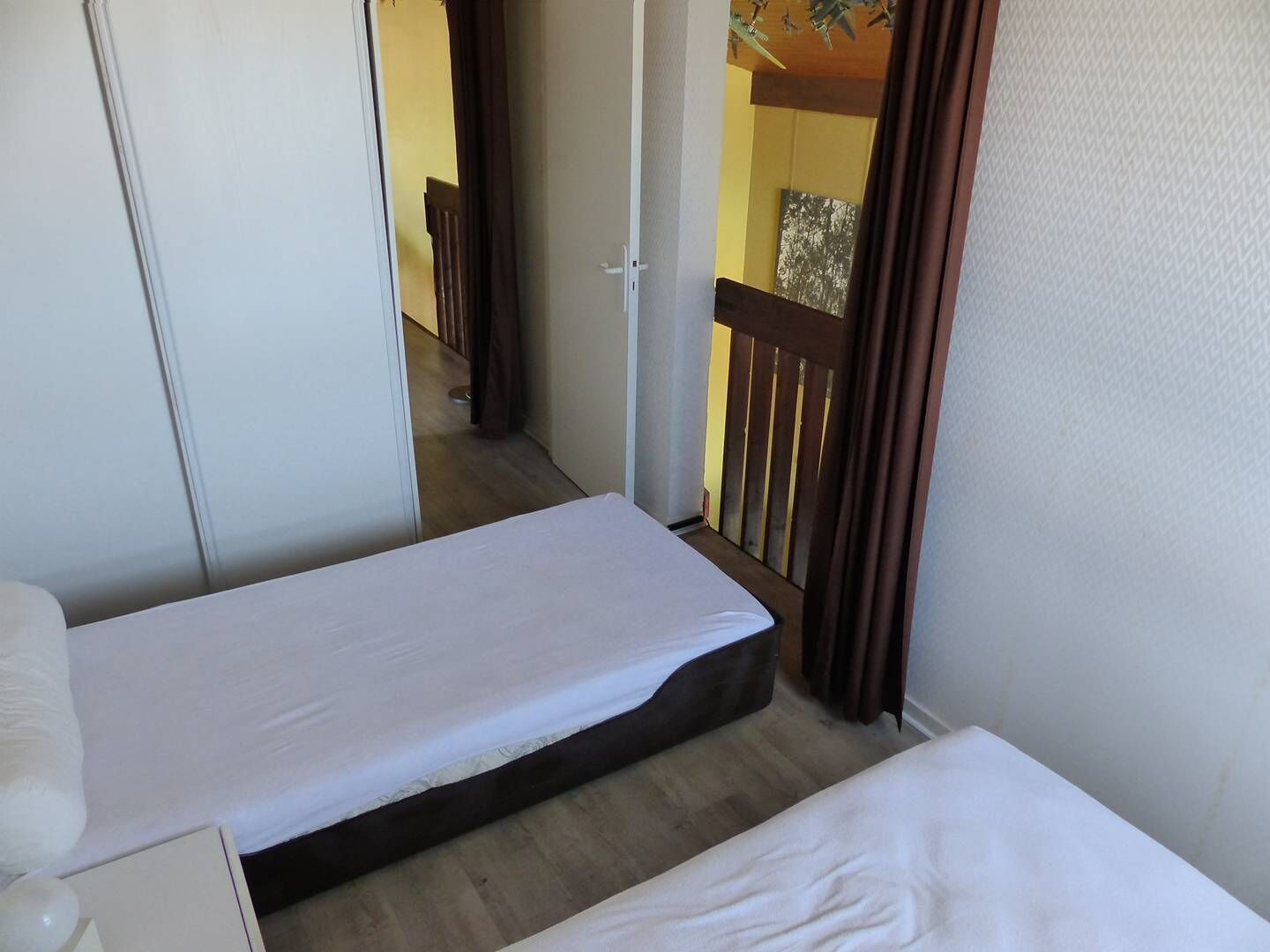 A small, narrow room with two single beds, a wooden floor, and a view of a balcony with a railing.