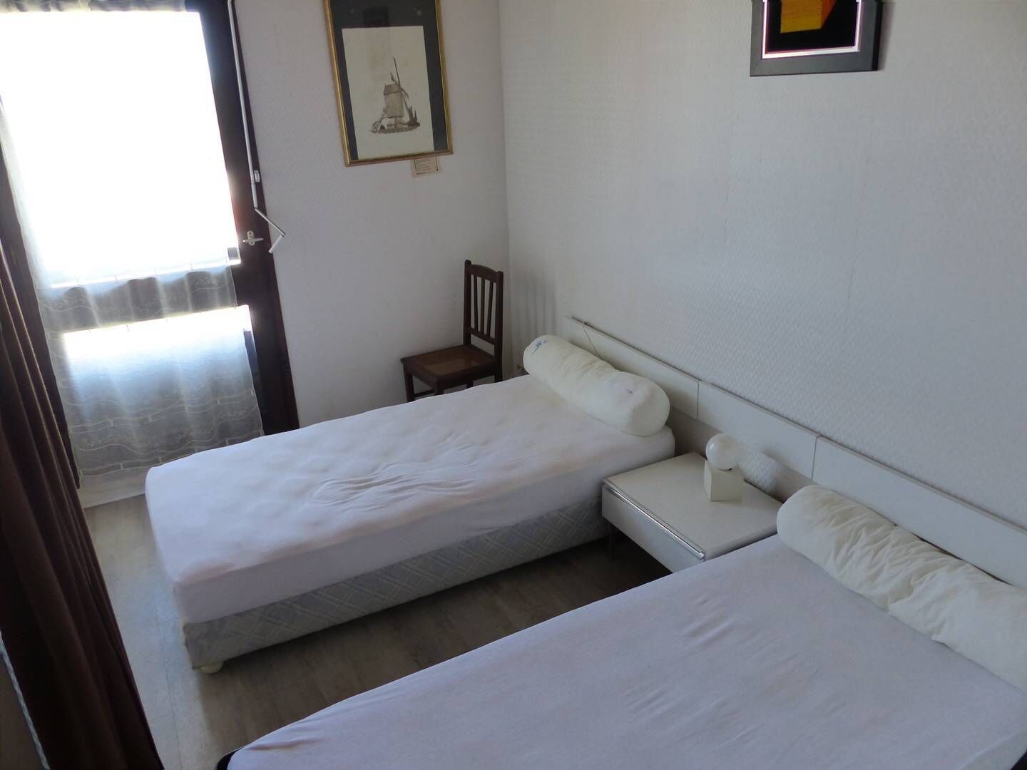A bedroom with two beds, a chair, a framed picture on the wall, and a window with curtains.