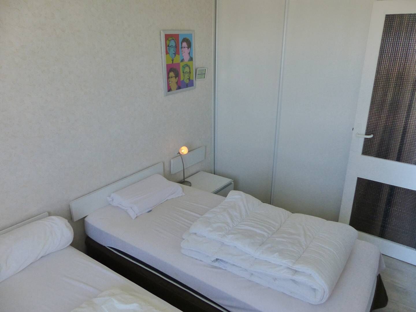 A hotel room with two beds, a bedside table with a lamp, a door with a mesh screen, and a wall with a poster.