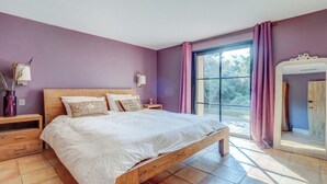 A bedroom with a wooden bed, purple walls, a large window with curtains, a nightstand, and a mirror. - Villa Tahiti (Ramatuelle)