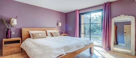 Un dormitorio con cama de madera, paredes moradas, un ventanal con cortinas, una mesita de noche y un espejo.