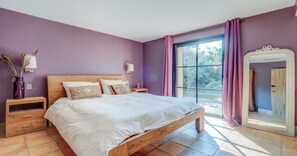 Ein Schlafzimmer mit einem hölzernen Bett, violetten Wänden, einem großen Fenster mit Vorhängen, einem Nachttisch und einem Spiegel.