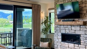 A modern living room with a stone fireplace, a flat-screen TV, and a large window overlooking a scenic landscape. - Lakeside Luxury: Hottub+Sauna+Fire Pit+Kid Friendly+Lake View+Mountain View (Whitefish)
