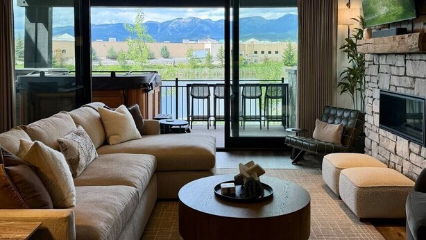 A living room with a sofa, ottoman, coffee table, and a fireplace. There is a balcony with a view of mountains and greenery. - Lakeside Luxury: Hottub+Sauna+Fire Pit+Kid Friendly+Lake View+Mountain View (Whitefish)