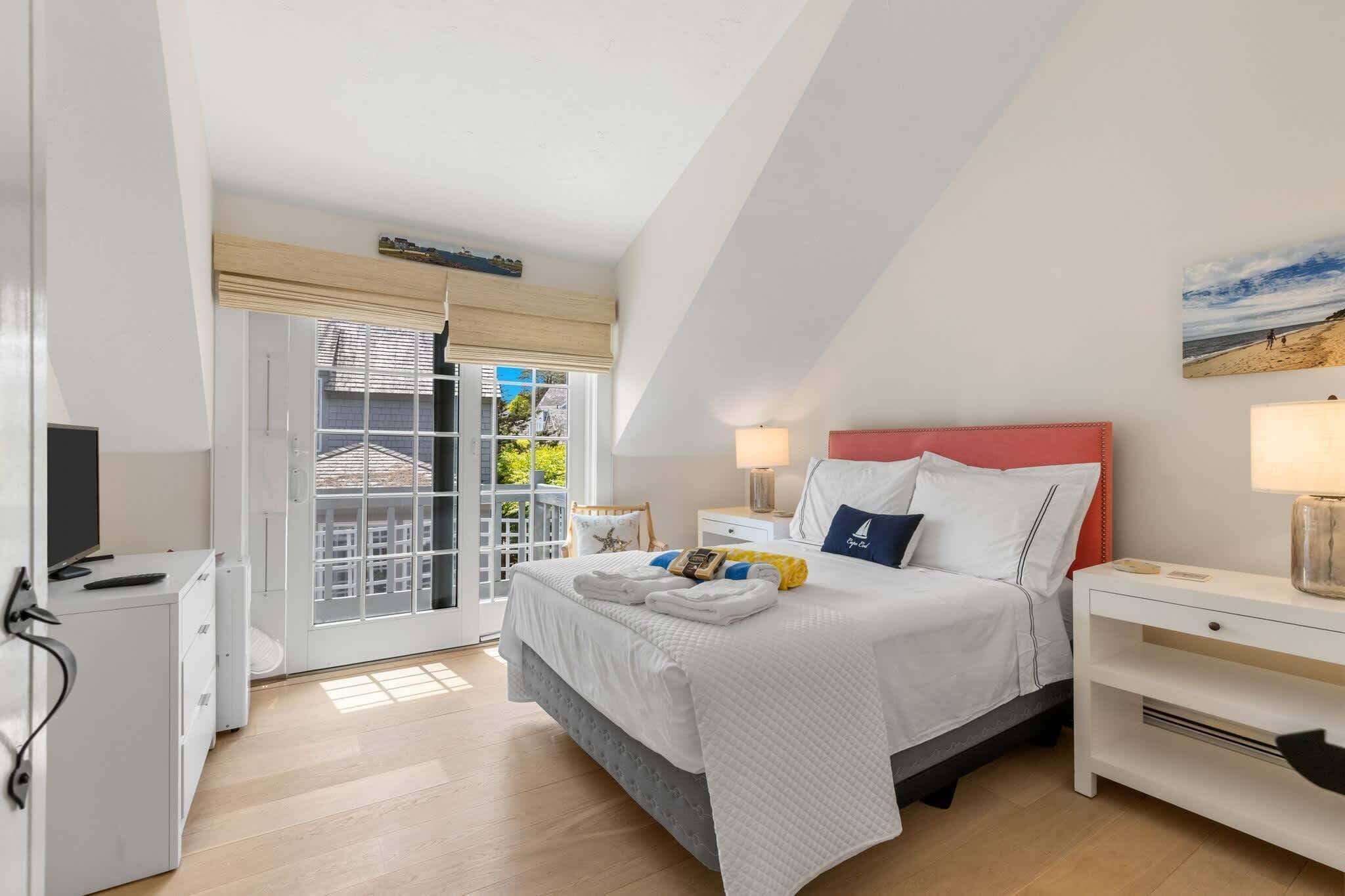 Un dormitorio con cama, mesitas de noche, televisor y puerta de cristal corredera que da a un balcón.