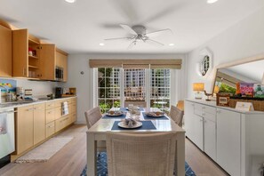A modern kitchen with wooden cabinets, a dining table set for two, and a view through a glass door to an outdoor area. - Hydrangea Lane Beach Cottage (Mashpee)