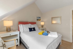 A bedroom with a bed, two bedside tables, a nightstand, a chair, and a door. - Hydrangea Lane Beach Cottage (Mashpee)