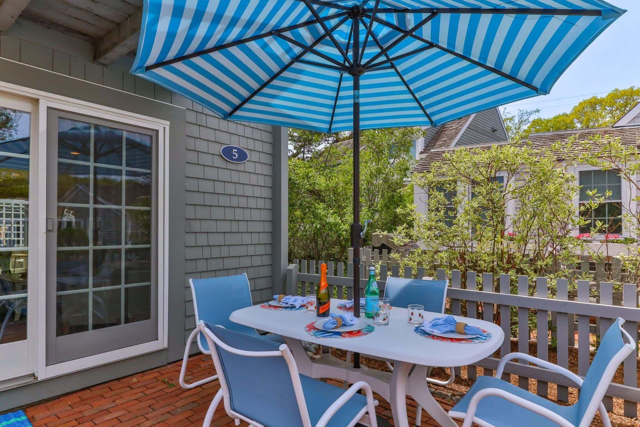 An outdoor patio with a table and chairs, a striped umbrella, and a white fence.