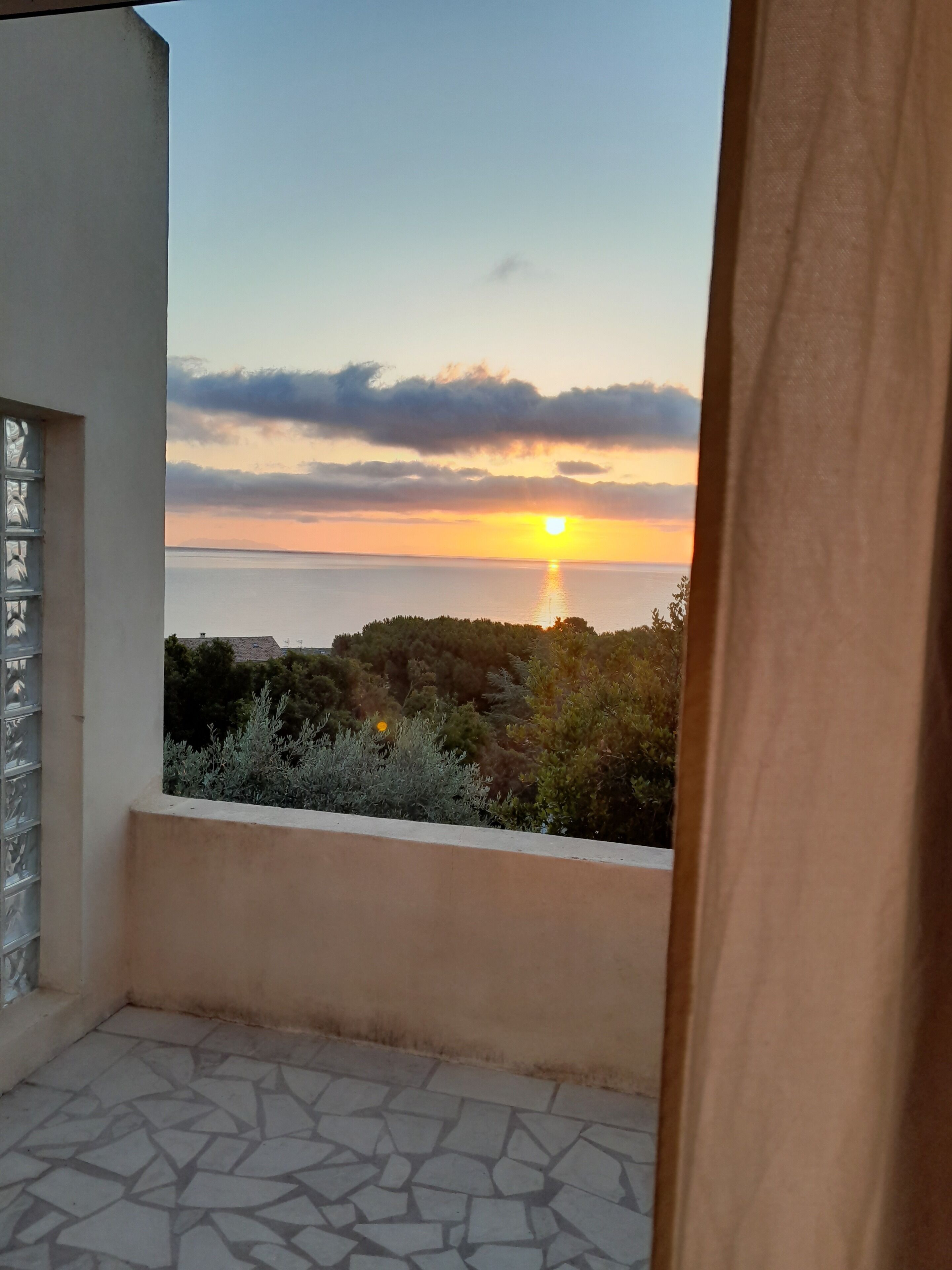 A balcony with a view of the sunset over the sea, stone flooring, and a white wall with a window.