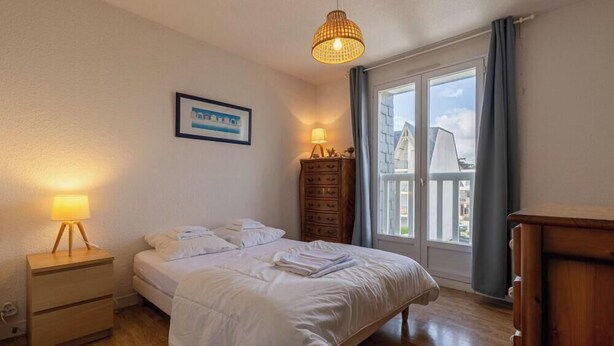 A bedroom with a bed, bedside table, lamp, dresser, and a window with curtains. - T3 apartment near the beach in Fréhel | 4 guests | Ref 117 (FREHEL)