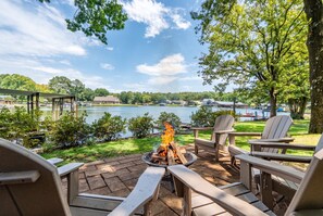 Eine Terrasse mit Adirondack-Stühlen, einem Feuerkorb und Blick auf einen See sowie Gebäude.