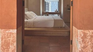 Ein schmaler Flur, der zu einem Schlafzimmer mit Bett, Fenster mit Metallgitter und Nachttisch mit Lampe führt.