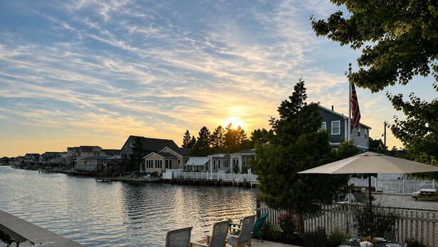Une terrasse au bord de l’eau, aménagée avec du mobilier d’extérieur et une table à manger, offrant une vue sur les maisons de l’autre côté.