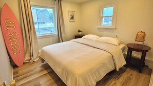 Une chambre à coucher avec un lit, deux fenêtres, une table de chevet avec une lampe, une petite chaise et un élément de décoration représentant une planche de surf.