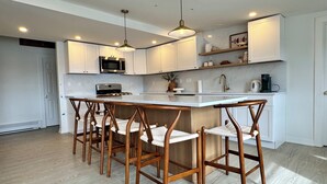 Une cuisine moderne avec des meubles blancs, un îlot central, des chaises en bois et des luminaires suspendus.