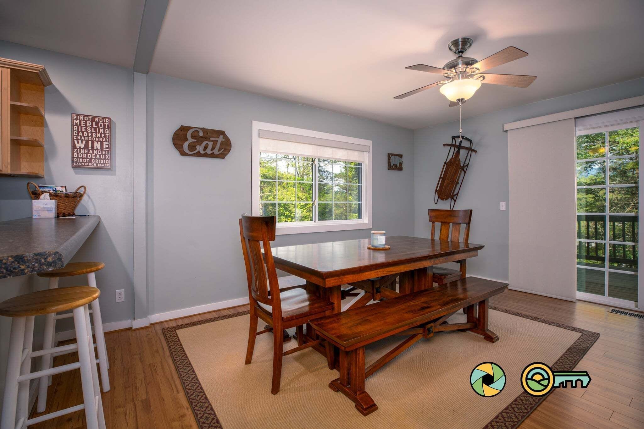 Un coin repas avec une table et des chaises en bois, un ventilateur de plafond et une fenêtre donnant sur un paysage verdoyant.