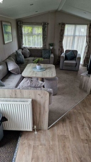 A cozy living room with a sofa, armchair, and coffee table, a radiator, and a window with curtains. - Southview Holiday Park - Holiday Accommodation 21279 (Skegness)