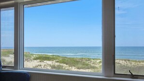 Ein Zimmer mit großem Fenster und Blick auf einen Strand mit Dünen und dem Meer.