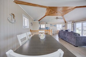 A spacious dining area with a long table and white chairs, a kitchen in the background, and a grey sofa in the living area. - 1725 - Sunshine and Whiskey 4BR oceanside retreat with private pool, hot tub, and deck access. (Corolla)
