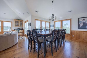 A spacious dining area with a large wooden table and chairs, surrounded by large windows offering views of the outdoors. - 1925 - Lemon Cooler Your Corolla dream stay: 6BR, pool, hot tub, theater room & sunny decks. (Corolla)
