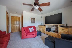 A living room with a ceiling fan, a large flat-screen TV, a red sofa, and wooden cabinets. - 1925 - Lemon Cooler Your Corolla dream stay: 6BR, pool, hot tub, theater room & sunny decks. (Corolla)