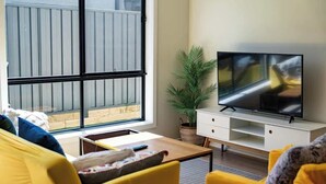Ein Wohnzimmer mit großem Fenster, Fernseher, gelben Sofas und einem Topfpflanzen.
