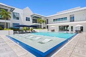 A swimming pool with lounge chairs and umbrellas, surrounded by a modern building with balconies and windows. - Olde Naples 2BR condo with pool view, heated pool, balcony, and walkable location. (Naples)