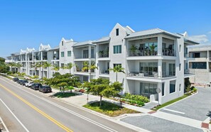 A row of modern multi-story residential buildings with balconies, a clear sky, and a street with parked cars. - Olde Naples 2BR condo with pool view, heated pool, balcony, and walkable location. (Naples)