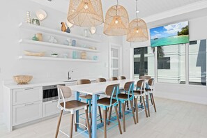 A modern kitchen with white cabinetry, a dining area with blue chairs, and a large window with a view of the ocean. - Olde Naples 2BR condo with pool view, heated pool, balcony, and walkable location. (Naples)
